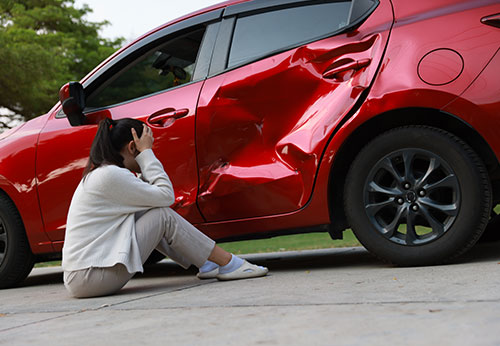Distressed woman sitting beside a red car with significant dent damage after a car accident